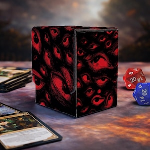 Puede incluir: Una caja negra para guardar cartas cubierta con un patrón repetido de ojos rojos. La caja está abierta, revelando el interior. Dos dados de múltiples caras son visibles en el fondo, junto con algunas cartas coleccionables.