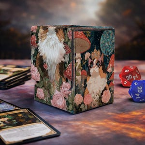 May include: A square card storage box with a whimsical design featuring a cat wearing a crown and glasses, surrounded by mushrooms and roses. The box is decorated with a forest theme. Two dice and playing cards are in the foreground.