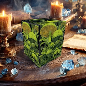 Op de afbeelding: Een zwarte kubusvormige doos met een groen paddenstoel- en schedelontwerp. De doos wordt omringd door kaarsen, kristallen en oude boeken. De paddenstoelen hebben een geelgroene kleur en de schedels zijn zichtbaar.