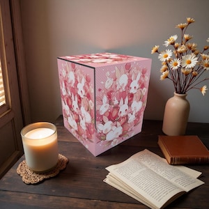 May include: A pink cube lamp featuring a pattern of white rabbits and red flowers. The lamp sits on a dark wooden surface, alongside a lit candle, a vase of flowers, and an open book.