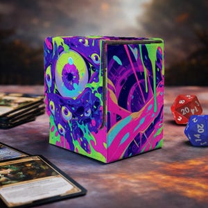 Op de afbeelding: Een kubusvormige kaartenbox met een levendig, psychedelisch ontwerp met meerdere ogen en kleurrijke druppels in neon groen, paars en roze. De doos staat naast speelkaarten en dobbelstenen.