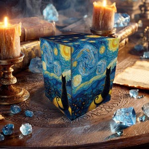 Op de afbeelding: Een kubusvormig decoratief item met een sterrennachtontwerp en zwarte kat silhouetten. Het kunstwerk is in de stijl van Van Gogh, met wervelende blauwe, gele en donkere accenten. Aangestoken kaarsen en kristallen omringen de kubus op een houten oppervlak.