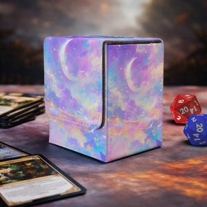 May include: A square card storage box with a celestial design featuring a crescent moon, stars, and clouds in shades of purple, blue, and pink. The box has a magnetic closure and is next to a red and blue die.