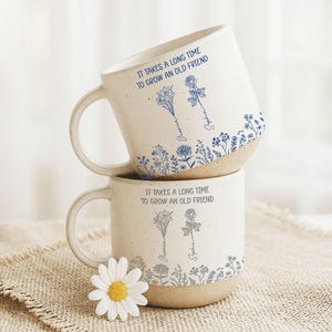Könnte beinhalten: Zwei gestapelte weiße Keramikbecher mit blauen Blumendesigns und dem Text "IT TAKES A LONG TIME TO GROW AN OLD FRIEND." Jeder Becher hat einen Henkel und einen beigefarbenen Boden. Eine Gänseblümchen ist im Vordergrund.