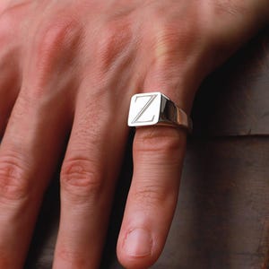 May include: A silver signet ring with the letter "Z" engraved on the square face. The ring is worn on a finger, showcasing its polished metal finish. The ring is a classic piece of jewelry.