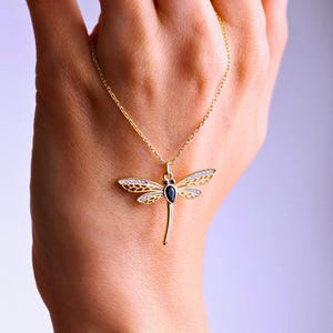 May include: A gold-colored dragonfly pendant necklace. The dragonfly has openwork wings with clear crystals and a dark blue center stone. The necklace is on a delicate gold chain.
