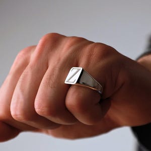 Op de afbeelding: Een zilveren zegelring met de letter "Z" gegraveerd op een rechthoekig vlak. De ring wordt aan een vinger gedragen, waarbij het gepolijste metaal en het eenvoudige ontwerp worden getoond. De achtergrond is zachtgrijs.