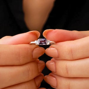 May include: A silver ring with a rectangular, deep purple gemstone at the center, flanked by two smaller, clear stones. The ring is held between two hands against a dark background.