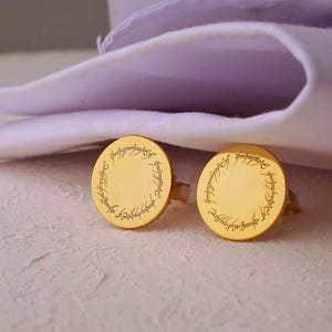Könnte beinhalten: Zwei goldfarbene Manschettenknöpfe mit kreisförmigem Design und eingraviertem Text. Sie liegen auf einer hellen Oberfläche, mit einem gefalteten lila Stoff im Hintergrund. Das Design erinnert an ein Fantasy-Thema.