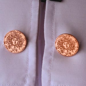 May include: Two copper-colored cufflinks, each featuring a detailed face with flowing hair, are displayed on a white shirt. The cufflinks are round and have an antique or vintage aesthetic. The shirt's fabric is smooth and light.