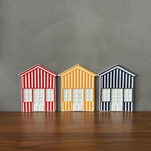 Peut inclure: Trois ornements décoratifs plats en forme de maison avec des rayures verticales rouges, jaunes et bleu foncé. Chaque ornement présente une porte centrale blanche et deux fenêtres blanches à carreaux, sur un fond de couleur neutre.