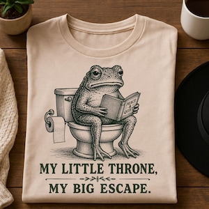 May include: A beige t-shirt featuring a dark green graphic of a toad sitting on a toilet reading a book titled Peace & Quiet. Below the graphic, the text reads: MY LITTLE THRONE, MY BIG ESCAPE. The design includes a toilet paper roll holder.