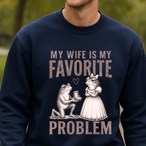 May include: A navy blue crewneck sweatshirt featuring a graphic print of a frog kneeling to propose to another frog wearing a dress. The text reads MY WIFE IS MY FAVORITE PROBLEM, with a small heart icon positioned between the words FAVORITE and the frogs.