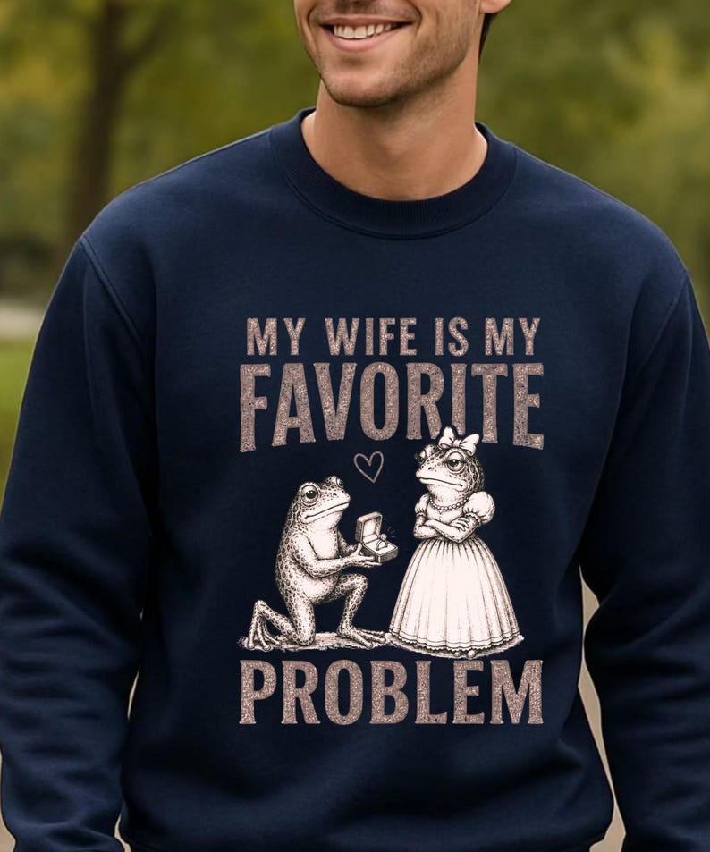 May include: A navy blue crewneck sweatshirt featuring a graphic print of a frog kneeling to propose to another frog wearing a dress. The text reads MY WIFE IS MY FAVORITE PROBLEM, with a small heart icon positioned between the words FAVORITE and the frogs.