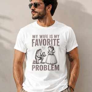 May include: A white short-sleeved t-shirt featuring a graphic of a frog kneeling to propose to another frog wearing a dress. The text reads MY WIFE IS MY FAVORITE PROBLEM with a small heart icon between the words. The design is in a dark brown tone.