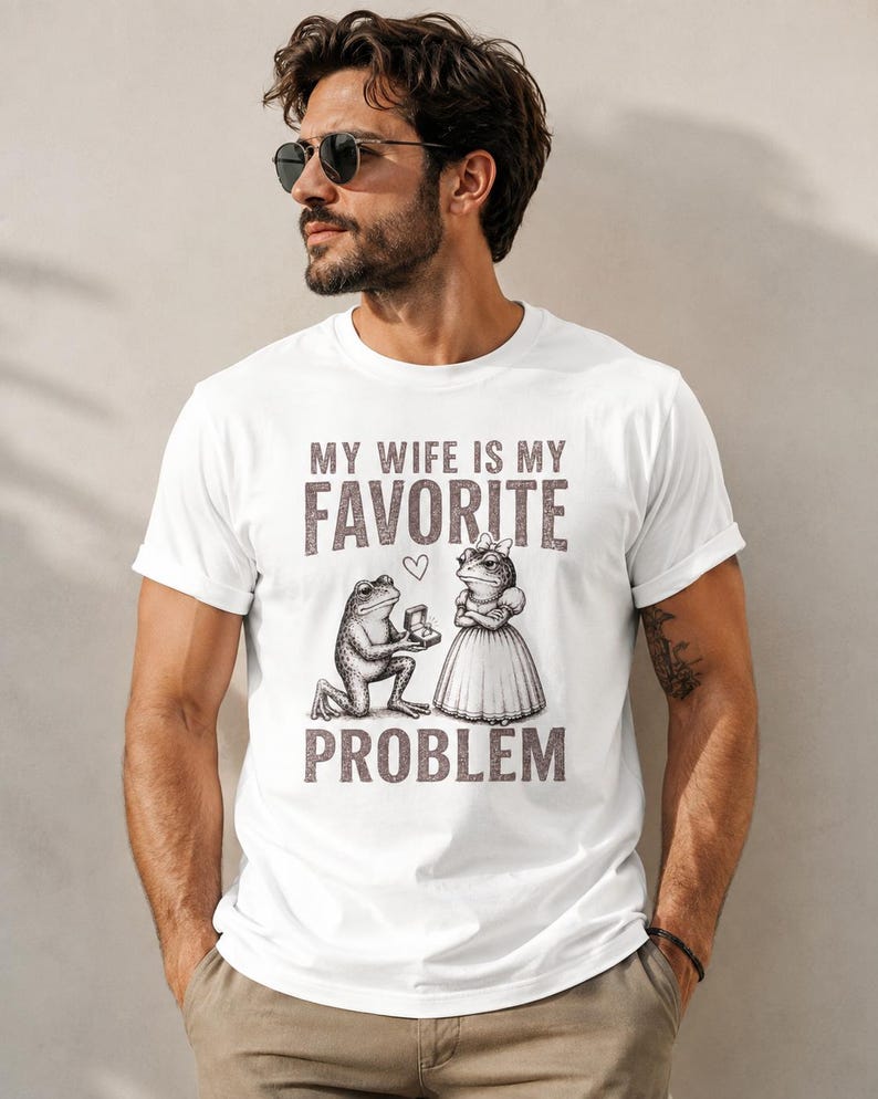 May include: A white short-sleeved t-shirt featuring a graphic of a frog kneeling to propose to another frog wearing a dress. The text reads MY WIFE IS MY FAVORITE PROBLEM with a small heart icon between the words. The design is in a dark brown tone.