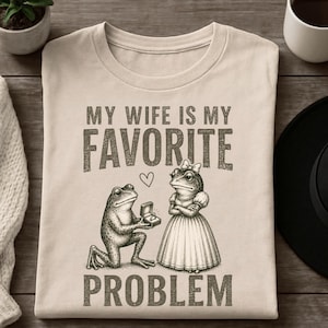 May include: A beige t-shirt featuring a dark brown graphic of a frog kneeling and offering a ring box to a frog wearing a dress and a bow. Above and below the illustration, the text reads: MY WIFE IS MY FAVORITE PROBLEM, with a small heart.
