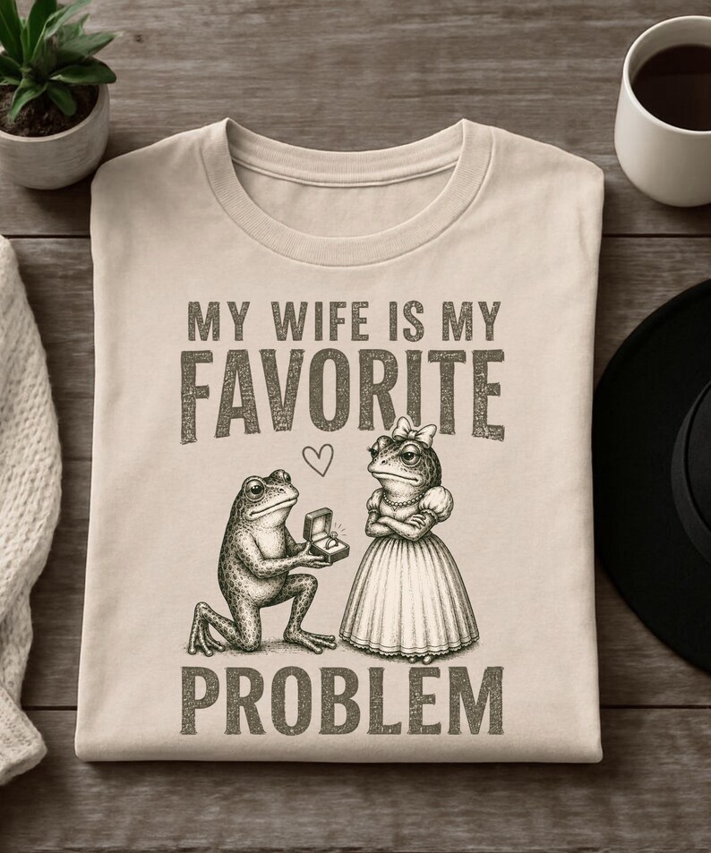 May include: A beige t-shirt featuring a dark brown graphic of a frog kneeling and offering a ring box to a frog wearing a dress and a bow. Above and below the illustration, the text reads: MY WIFE IS MY FAVORITE PROBLEM, with a small heart.