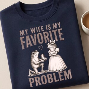 May include: A navy blue crewneck sweatshirt featuring a graphic of two frogs. One frog kneels, offering a ring box to the other frog, who wears a dress and bow. The text reads "MY WIFE IS MY FAVORITE PROBLEM" in a textured, light-colored font.