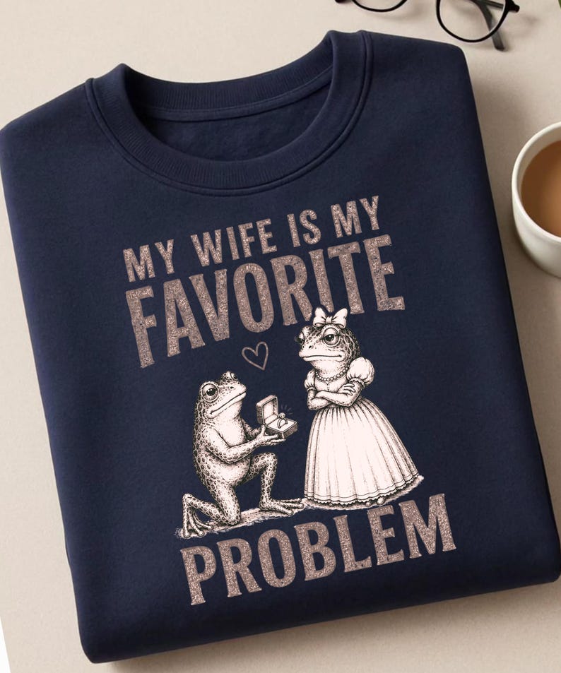May include: A navy blue crewneck sweatshirt featuring a graphic of two frogs. One frog kneels, offering a ring box to the other frog, who wears a dress and bow. The text reads "MY WIFE IS MY FAVORITE PROBLEM" in a textured, light-colored font.