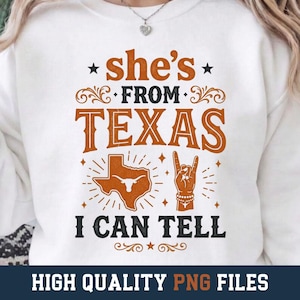 Może przedstawiać: Biała bluza z okrągłym dekoltem i pomarańczowo-czarnym nadrukiem graficznym. Tekst brzmi "she's FROM TEXAS I CAN TELL", a obok znajduje się sylwetka stanu Teksas z ikoną longhorna oraz dłoń wykonująca gest rock-and-roll.