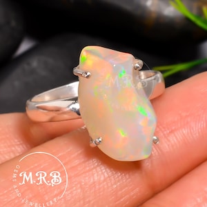Peut inclure: Bague en argent ornée d'une opale brute de grande taille. L'opale présente des couleurs blanc crème, jaune et vert. Le corps de la bague est en argent poli. La pierre est maintenue par quatre griffes en argent. Bijou unique.