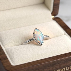 May include: A marquise-cut opal ring with a silver band, presented in a dark wood jewelry box with a cream-colored interior. The box features the text M.R.B GEMS AND JEWELLERY engraved on the front and a circular logo with the same text.
