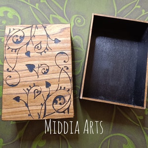 Op de afbeelding: Een houten doos met een zwart interieur en een bloemmotief dat in de deksel is gegraveerd. De doos is gesloten en de deksel is gericht op de camera. De achtergrond is een groene en bruine gepatenteerde stof.