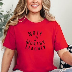 May include: A red t-shirt with the words "NOTE WORTHY TEACHER" in black script, with a musical note symbol. The shirt is short-sleeved and appears to be made of a soft material.