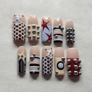 May include: A set of ten press-on nails featuring various designs including brown polka dots, silver metallic studs, red and blue stars, horizontal stripes, and solid red or beige bases. Each nail has a nude-colored base near the cuticle area.