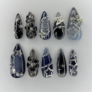 May include: A set of ten almond-shaped press-on nails in shades of dark blue and black. The nails feature silver-toned embellishments including star charms, butterfly accents, floral relief patterns, swirling lines, and small metallic beads.