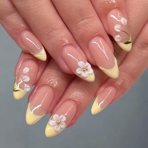 May include: Almond-shaped nails featuring a nude base with pale yellow French tips. Some nails are adorned with three-dimensional white floral accents with gold centers, while others feature delicate painted white flowers with green leaves.