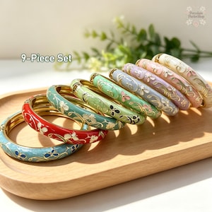 May include: A 9-piece set of round bangles featuring gold-tone metal edges and enamel surfaces. Each bangle displays a floral and butterfly motif in various colors including red, light blue, green, lavender, pink, and cream with gold accents.
