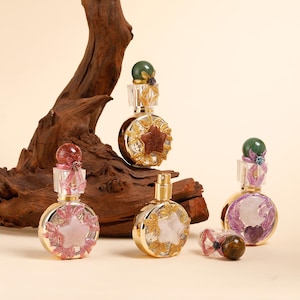 Puede incluir: Una variedad de frascos de perfume decorativos con detalles dorados y diseños en forma de estrella. Los frascos presentan varias piedras preciosas de colores y adornos florales, con un fondo de madera.