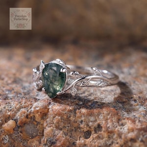 Könnte beinhalten: Ein silberner Ring mit einem birnenförmigen grünen Moosachat-Edelstein. Der Ring hat ein zartes Blattdesign am Band und um den Edelstein herum. Der Ring ist vor einem strukturierten, braunen Hintergrund platziert. Der Text "Porcelain ParlorShop" befindet sich oben links.