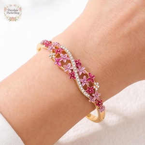 May include: Gold-toned bracelet with a floral design. The bracelet features pink and purple flowers and a row of clear crystals. The bracelet is worn on a wrist. The brand name "Porcelain Parlor Shop" is in the upper left corner.