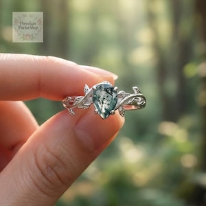 Può includere: Un anello d'argento con una gemma verde muschio a forma di pera. La fascia dell'anello è decorata con dettagli di foglie e viti. L'anello è tenuto su uno sfondo verde sfocato, che mette in risalto il suo design.