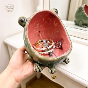 May include: A ceramic frog-shaped trinket dish with a pink interior and green exterior. The dish contains gold bracelets, a ring, and small decorative items. The Porcelain Parlor Shop logo is visible in the upper left corner.
