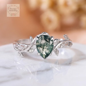 May include: A silver-toned ring featuring a pear-shaped moss agate gemstone with dark green inclusions. The band is designed with a detailed vine and leaf motif that wraps around the center stone, held in place by four small metal prongs.