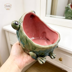 May include: A ceramic frog-shaped planter with a glossy green exterior and a pink interior. The frog has large, black eyes and detailed feet. The planter is likely for small plants or decorative items. The Porcelain Parlor Shop logo is in the upper left corner.