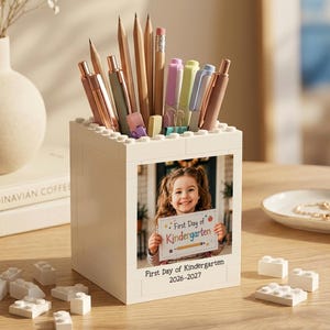 May include: A white square pencil holder constructed from building blocks, featuring a photo of a child holding a sign that reads First Day of Kindergarten and the text First Day of Kindergarten 2026-2027. The holder contains various pens and pencils.
