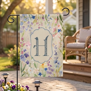 May include: A rectangular garden flag featuring a watercolour floral pattern with blue, pink, and yellow flowers on a cream background. A central scalloped frame contains a large, dark blue capital letter H with a decorative leaf motif.