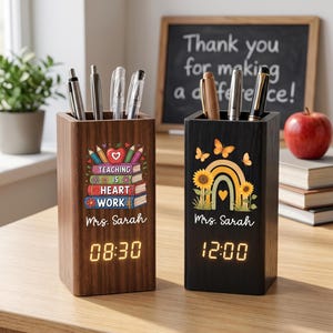 May include: Two rectangular wooden pen holders with digital clocks. The left holder is brown with a stack of books, an apple, and the text "Teaching is heart work" and "Mrs. Sarah". The right holder is black with a rainbow, sunflowers, and "Mrs. Sarah".