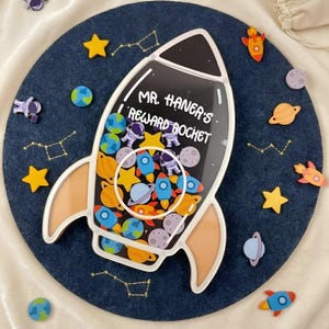 以下が含まれることがあります： 「MR. HANER'S REWARD ROCKET」という文字が書かれた透明な前面パネルを持つロケット型の報酬用ジャー。中には惑星、星、ロケット、宇宙飛行士などのカラフルな宇宙をテーマにしたトークンが入っており、濃い青色の円形の背景の上に配置されています。