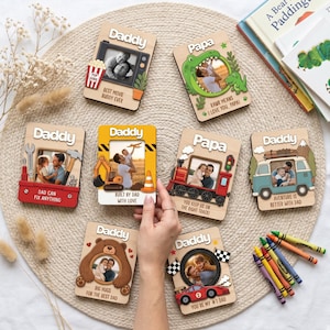 Op de afbeelding: Een verzameling houten fotolijsten met de woorden "Daddy" of "Papa" en verschillende ontwerpen. De lijsten hebben thema's als tv, dinosaurus, bouw, trein, beer en raceauto. Elke lijst bevat een foto.