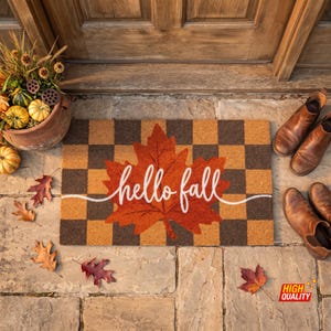 May include: A rectangular doormat featuring a brown and tan checkered pattern with a large orange maple leaf in the centre. The text "hello fall" is written in white cursive script across the leaf, extending horizontally across the mat.
