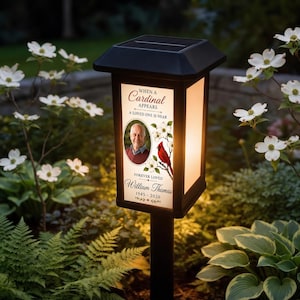 May include: A black solar-powered garden lantern featuring a memorial graphic with a portrait of a man, a red cardinal bird, white flowers, and the text: When a Cardinal Appears A Loved One Is Near Forever Loved William Thomas 1945-2026.