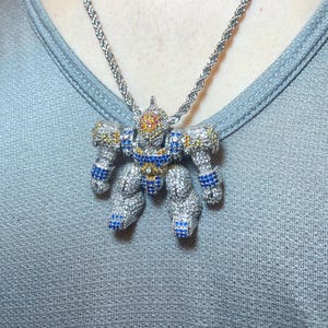 May include: A silver robot pendant necklace with a rope chain. The robot is encrusted with small, colorful gemstones in blue, yellow, and red. The necklace is worn on a gray shirt.