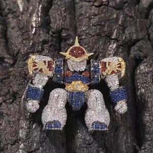 May include: A detailed, jewel-encrusted robot pendant. The robot features a silver body with blue, red, and gold accents. The pendant is covered in small, sparkling stones, creating a luxurious and eye-catching design. The robot's design is intricate and detailed.