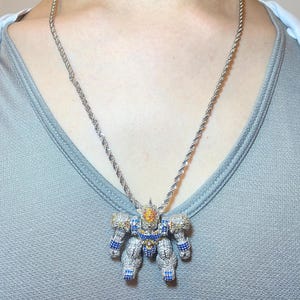 May include: Silver-toned necklace with a robot pendant. The pendant is encrusted with clear, blue, and yellow gemstones. The chain has a twisted rope design. The robot pendant has detailed arms, legs, and a head.